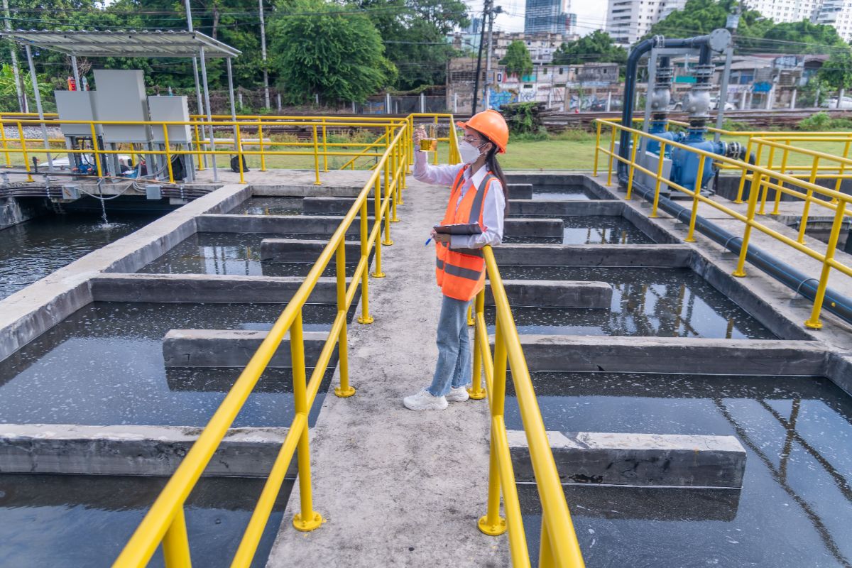 Water treatment technician in safety gear inspecting a water sample beside industrial treatment basins with yellow railings