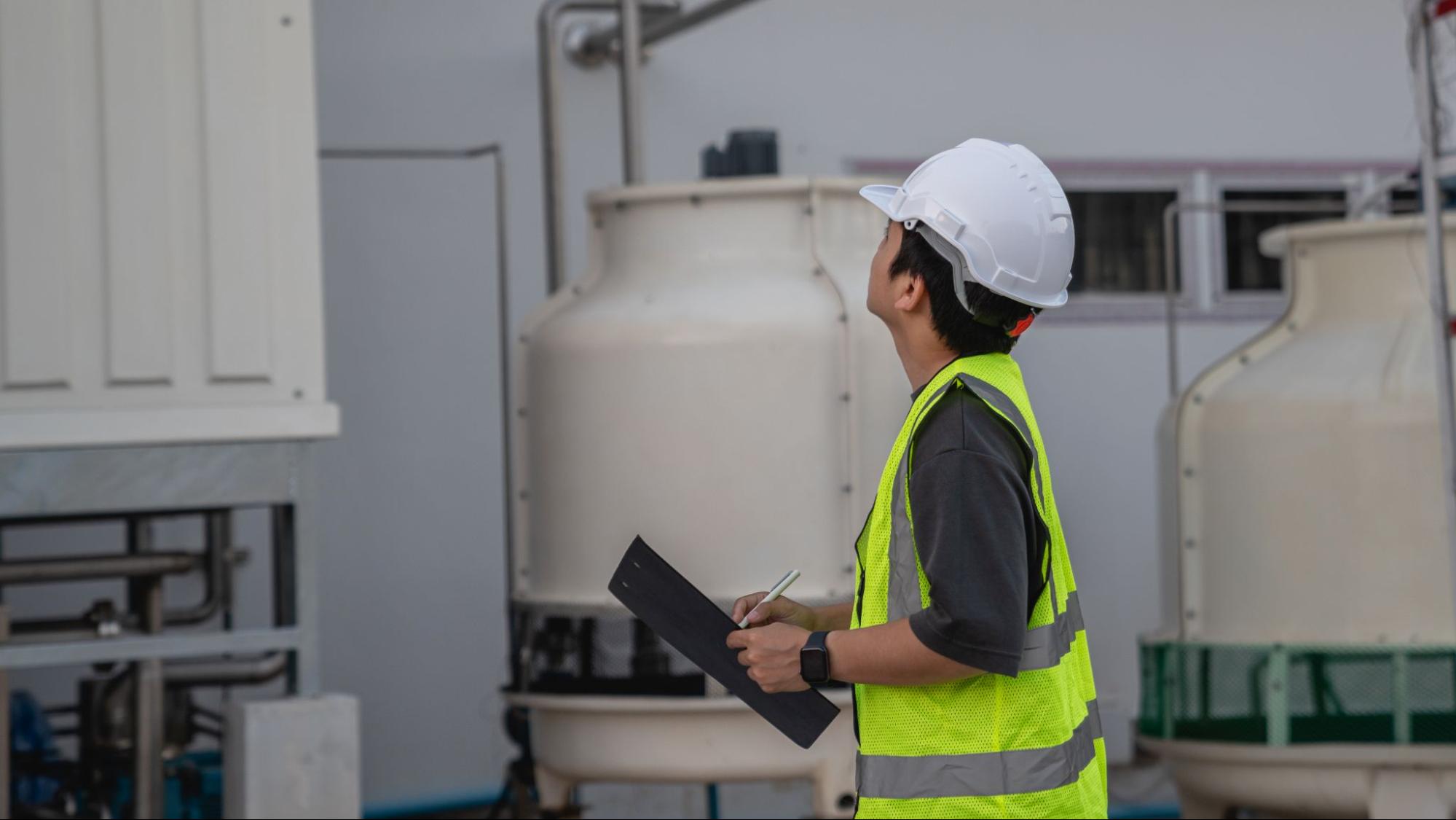 Technician in a hard hat and safety vest inspecting industrial water treatment equipment while holding a clipboard